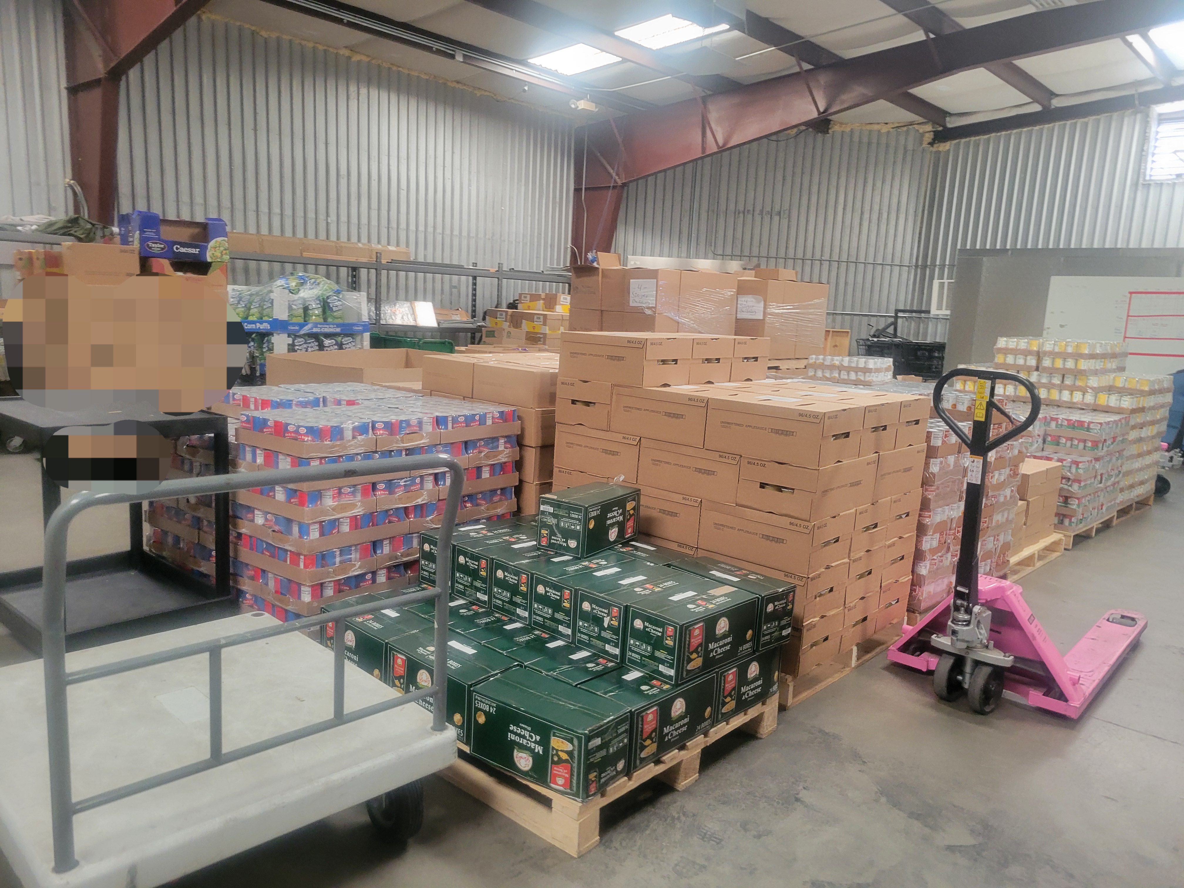 Pallets of food in a warehouse with shelf cart, pallet jack, and truck cart near by.